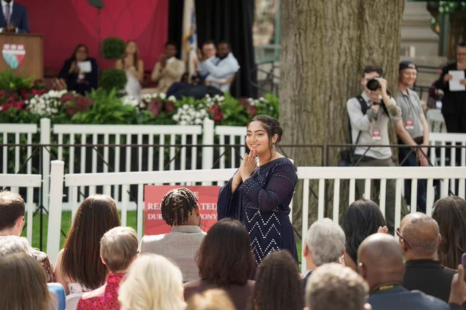 A person stands in Holmes Field, appearing joyful and clapping hands, with an audience and photographers in the background.