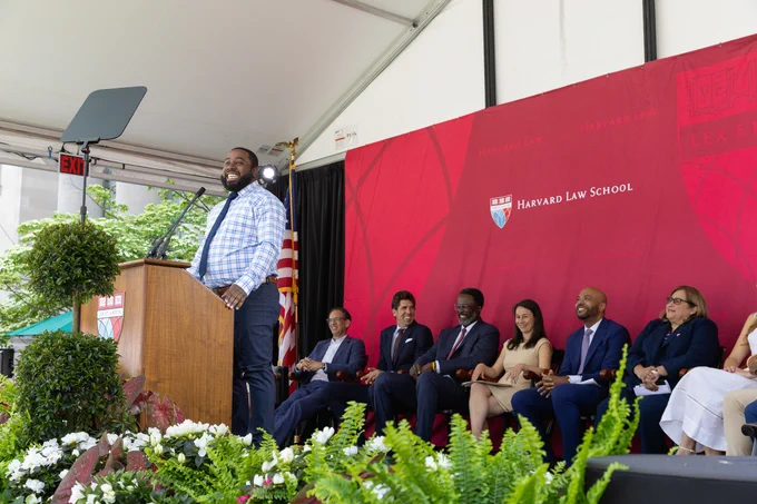 A person speaks at a podium during an outdoor event at Harvard Law School, with several other individuals seated behind them under a banner with the school's name.