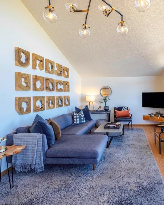 Interior of a living room featuring a gray sectional sofa with blue and gray pillows, a wooden coffee table, and modern lighting overhead. A wooden art installation adorns the white wall, and a flat-screen TV is mounted on the opposite wall.