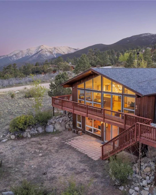 View of a warmly lit two-story cabin with large windows, situated in a serene landscape with distant views of snow-capped mountains.