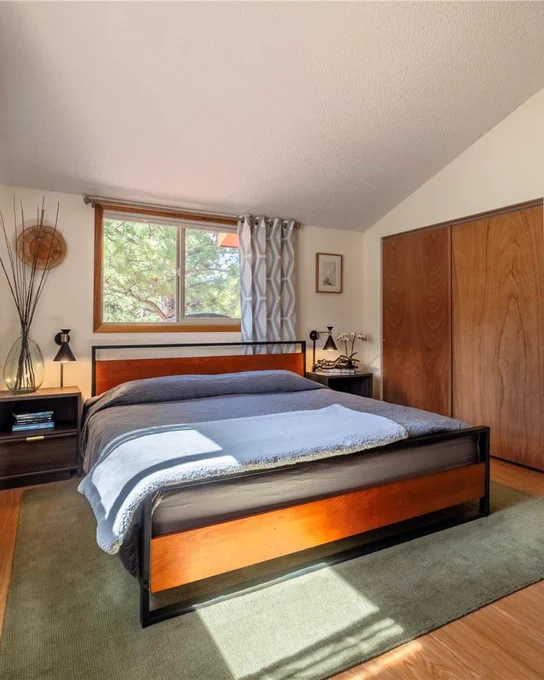 Interior of a well-lit bedroom featuring a large bed with a wooden frame, neatly made with a gray blanket. Beside the window with patterned curtains, there is a side table with a lamp and books. A spacious wooden closet is also visible. The room has a carpeted floor and a warm ambiance.