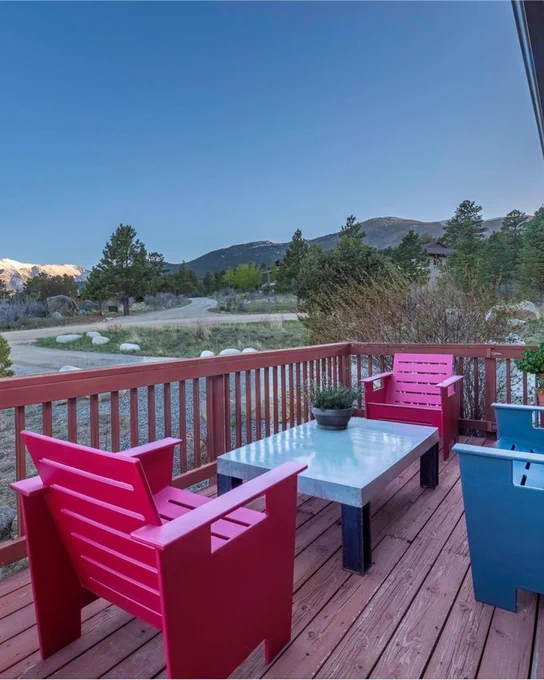 View from a balcony with red and blue chairs around a white table, overlooking a tranquil landscape with mountains and trees under a clear sky.