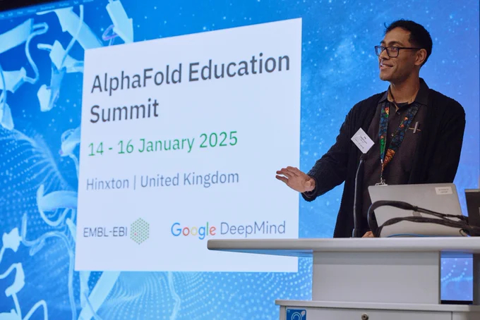 A man smiles at a lectern, talking towards a room. Behind him, a PowerPoint screen is shown with a blue background and the words 'AlphaFold Education Summit. 14 – 16 January 2025. Hinxton, United Kingdom. The EMBL's European Bioinformatics Institute and Google DeepMind logos are also shown on screen.