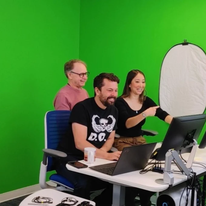 Three people stand smiling behind computer screens. The wall behind them is painting a bright green.