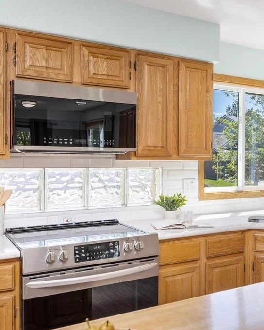A bright kitchen with wooden cabinets, a stainless steel stove, and a microwave above the stove. There is a window with a view of greenery.