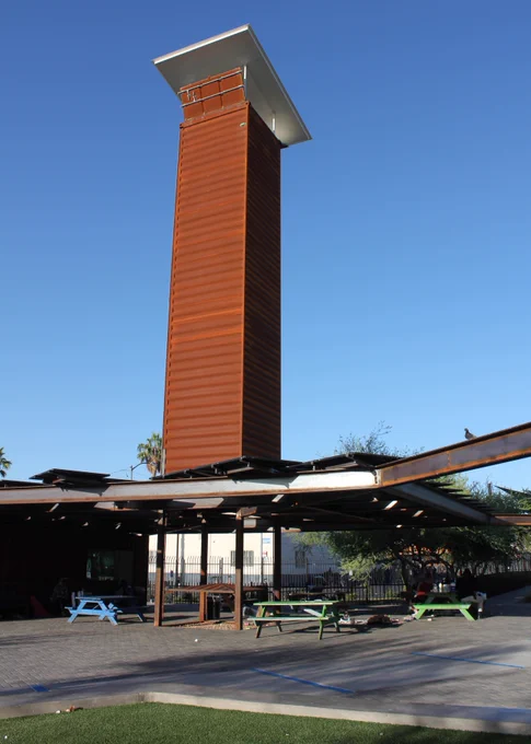 View of the tall, cooling tower, adjacent to an outdoor seating area with picnic tables and benches under a partially shaded structure. Clear blue sky in the background.