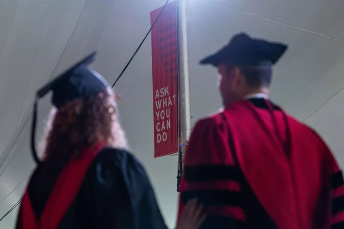 Two out of focus people standing next to each other in graduation regalia, with a in-focus sign between them that reads “Ask what you can do.”