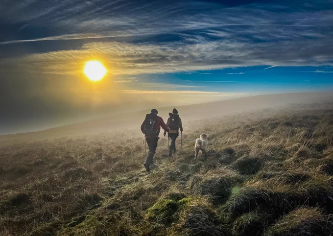 Zwei Menschen mit Wanderrucksäcken und ein Hund gehen bei tiefstehender Sonne durch nebliges, offenes Moorland. Die Szene wirkt atmosphärisch und weitläufig – ein früher Morgen oder später Nachmittag in der Natur.