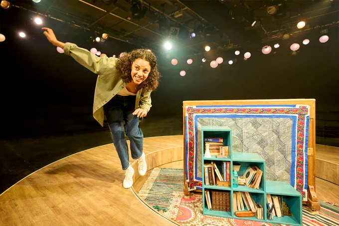 An actress on stage next to a small shelving unit packed with books.
