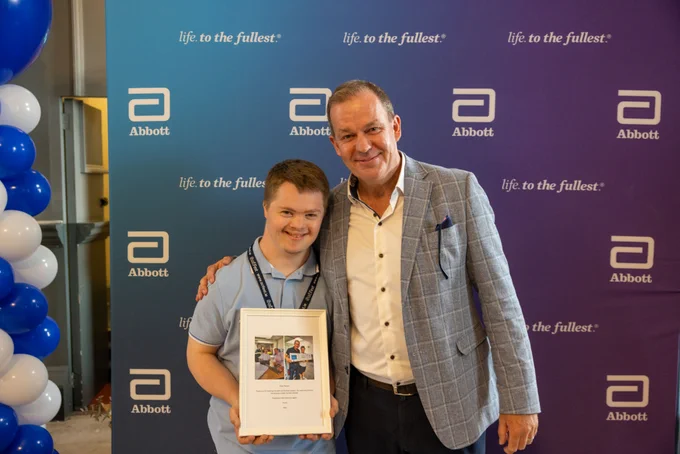 A TCPID student with his mentor from Abbott, holding a framed photo in front of an Abbott banner