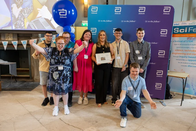 A group of TCPID students with one of the mentors from Abbott, who is holding a framed photograph with messages from each of the students. They are all standing in front of an Abbott banner