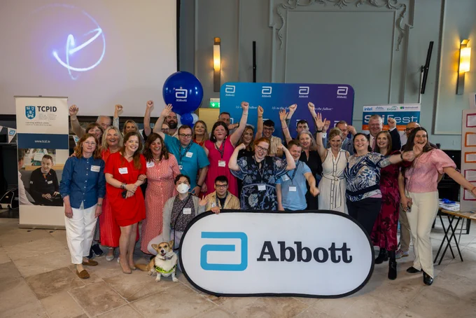A large group of TCPID students and staff with colleagues from Abbott cheering with arms raised and standing in front of an Abbott banner