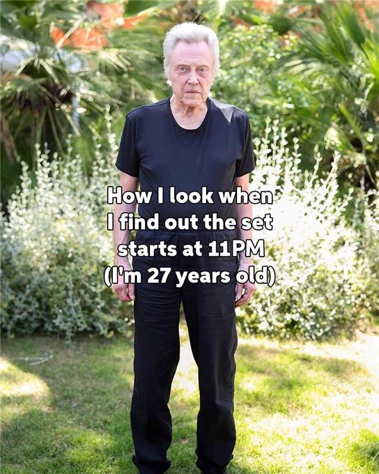 Christopher Walken stands in a garden, wearing a black t-shirt, with a caption expressing surprise about a late set start time, humorously noting a youthful age of 27 years.