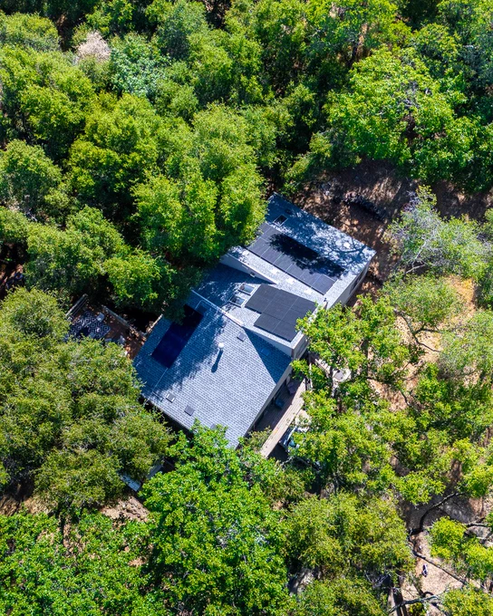 Aerial view of a modern house with solar panels on the roof, surrounded by dense green foliage.