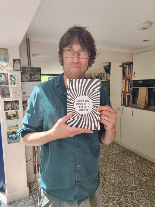 Here I am in my kitchen with a rather intense expression, displaying a proof of my book.  Its cover is black and white, with spiral patterns interrupted by a white circle in the centre, in which "CHOOSE YOUR PUZZLE-FILLED PATH TO TV STARDOM" is written.