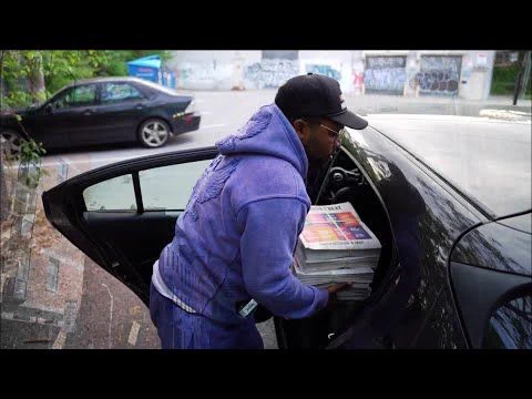 A man wearing a purple sweatshirt and sweatpants puts bundles of newspapers into a car. 
