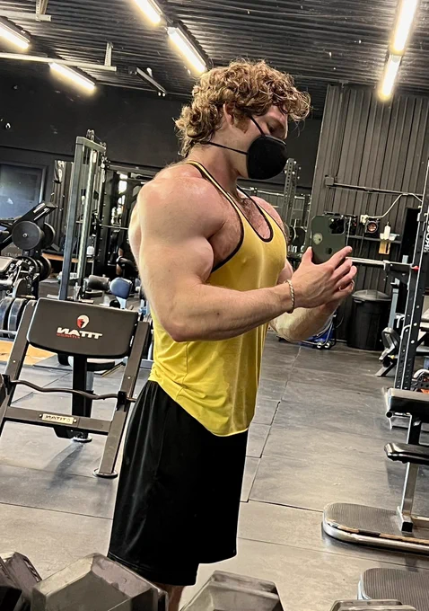 Gym mirror side profile photo of a hairy chested, muscular white man with an upper body pump. He’s wearing a yellow stringer tank top with black shorts, and a black Zimi Air KN100 respirator. He has medium length curly hair that is wet with sweat.