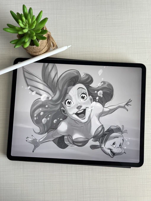 A digital illustration of Ariel and Flounder displayed on an iPad, with a stylus and a small potted plant beside it.