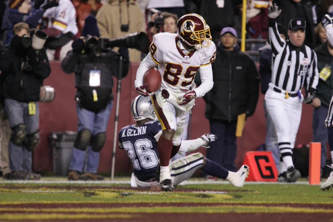 Photo of Santana Moss during a game. He is running in the end zone after scoring a touchdown. He is wearing a burgundy helmet, white jersey, white pants, burgundy socks, black cleats and burgundy football gloves.