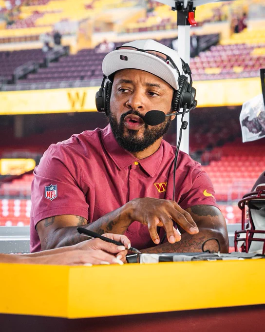 Photo of Santana Moss during a TV segment. He is sitting at a desk wearing headphones. He is also wearing a burgundy polo shirt and a white hat.