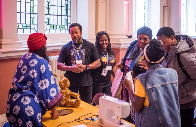 People chatting warmly around a sewing table in a brightly lit community hall with stained
glass windows. A sewing machine, handmade crafts, and cultural fabrics are on
display.