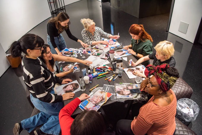 A diverse group of people sit and stand around a large table during a collage workshop. The
table is covered with art supplies, including markers, glue sticks, scissors, and magazines
with cut-out images. Participants are engaged in creating collages, some laughing and
sharing materials. The setting appears to be a modern, well-lit room with glossy black floors
and white walls.