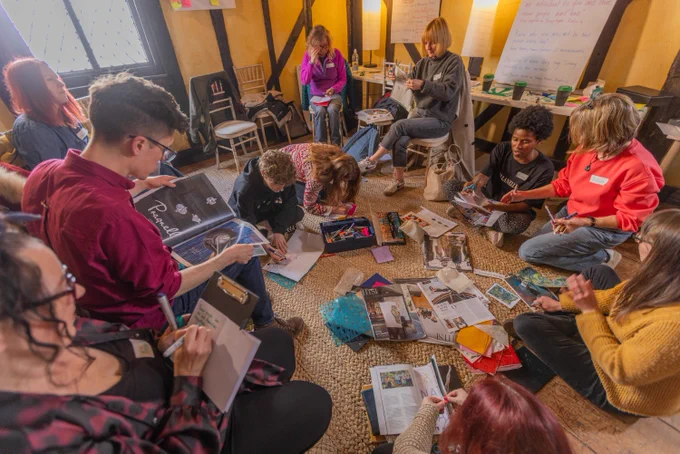 A diverse group of people gathered in a cozy, sunlit room, sitting in a circle on the floor and
chairs, engaging in creative activities with magazines, art supplies, and notebooks spread
out. 