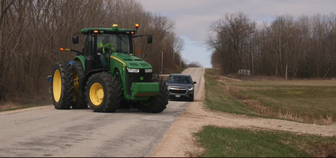 John Deere tractor driving on a rural road with a motorist waiting for it to turn