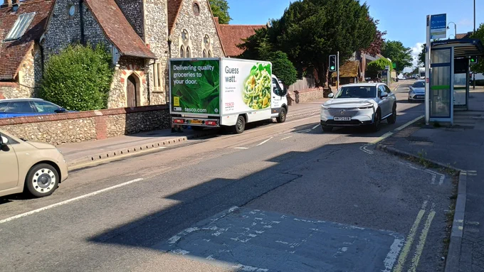 Picture of a tesco delivery can parked on zig zags at a traffic light controlled crossing outside a nursery. An empty layby can be seen less than 10m away. 