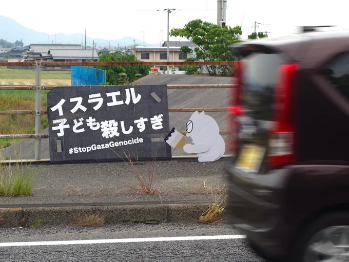 歩道の防護柵に立てかけられた「イスラエル子ども殺しすぎ」と書かれた大きなプラカード。隣に黒いインクのついたハケを持って怒る白いネコのイラストボード。車道を軽自動車が通り過ぎる。防護柵の向こうには畑、民家、山。