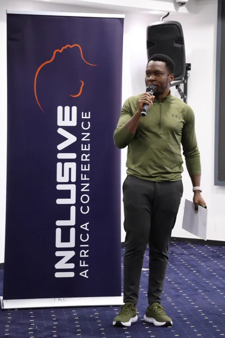 A man stands confidently speaking into a microphone next to a tall banner that reads "Inclusive Africa Conference." He is wearing a green long-sleeve athletic top, dark pants, and green sneakers, and is holding papers in his left hand. A speaker is mounted behind him on the wall in a modern conference room setting.