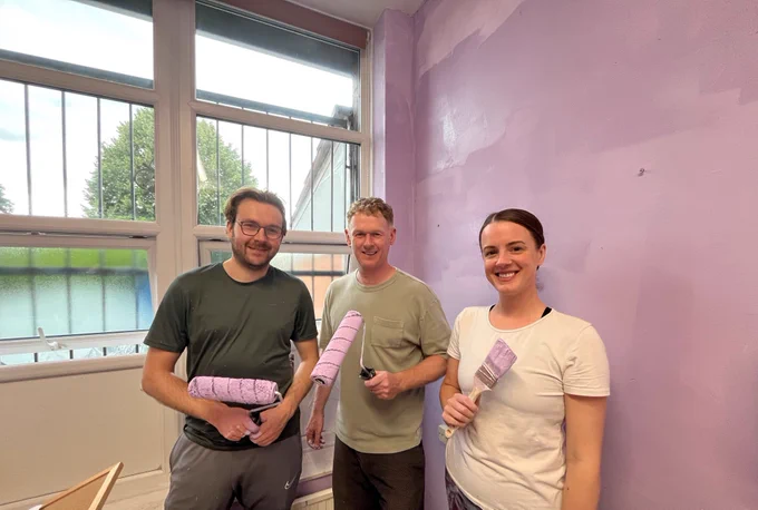 Two men and one woman stood in front of a newly painted lilac wall holding paint rollers and a paint brush. 