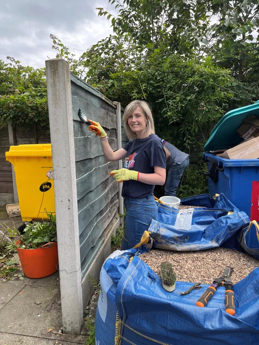 A lady with blonde hair wearing a blue t-shirt and jeans wearing yellow gloves painting a fence.