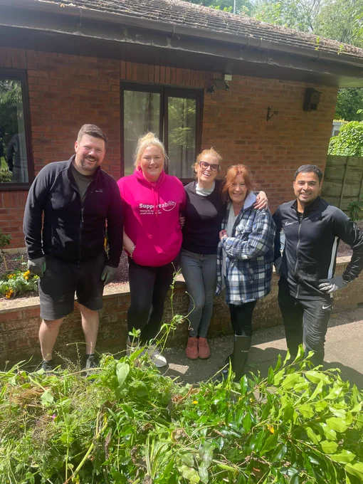 Three women and 2 men stood in a row in front of a bungalow and some green foliage in front of them. 