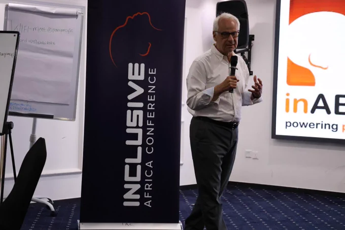 Wilfred Kainz from Zero Project stands next to an Inclusive Africa Conference banner, holding a microphone while presenting. He wears glasses, a white dress shirt, and dark trousers. A screen displaying the inABLE logo is visible in the background, along with a flipchart and part of the conference setup.