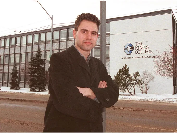 The photo shows Delwin Vriend standing outdoors in front of a building with a sign that reads "The King's College: A Christian Liberal Arts College." The individual is wearing a dark coat and has their arms crossed. The background includes a multi-story building with large windows, some evergreen trees, and a snowy landscape, suggesting a cold season. A streetlight pole and some bare trees are also visible.