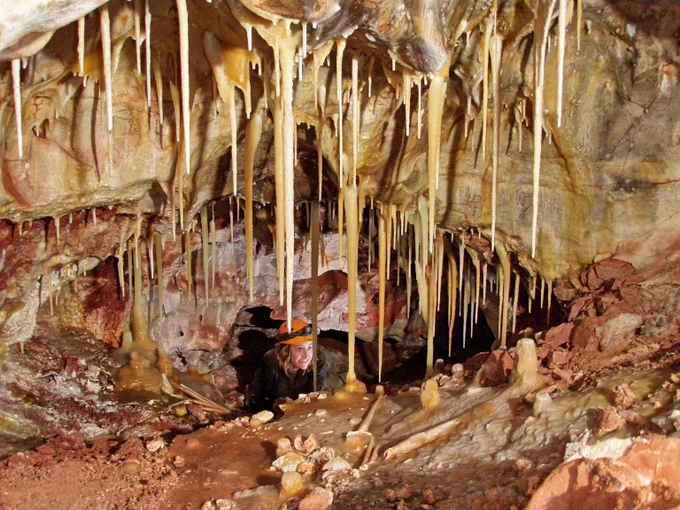 a smiling young woman in a helmet and headlamp looks up at stalactites in a cave