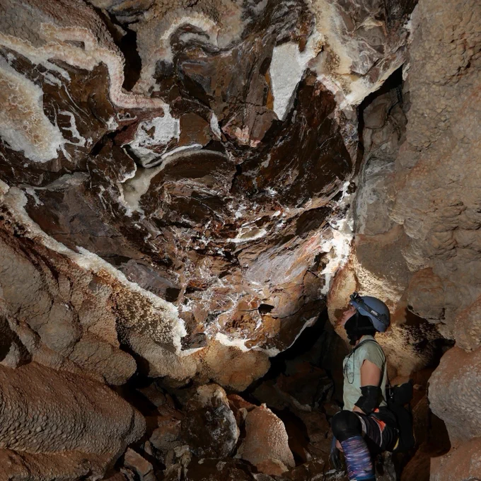 a caver in a helmet and headlamp looks up at cave formation above
