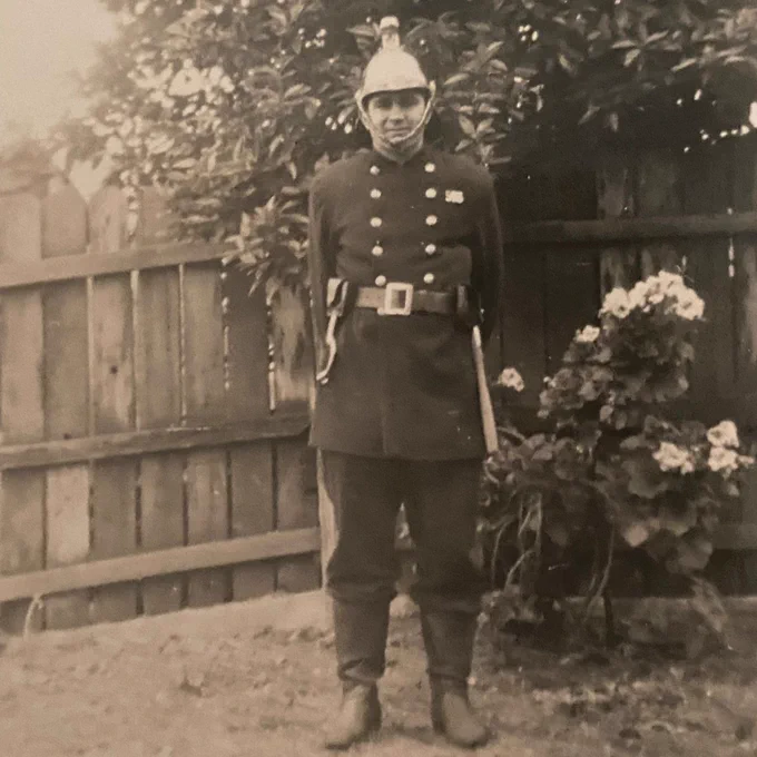 Harold as a firefighter, in his uniform standing beside a flowering bush against a wooden fence.
