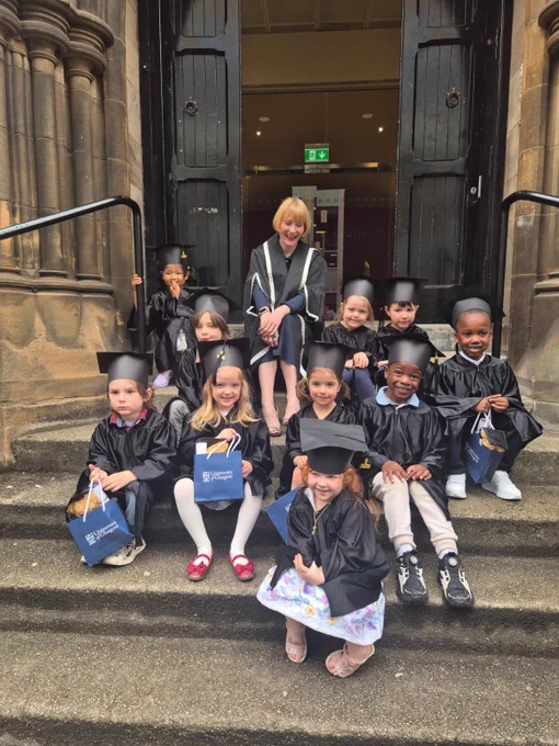 Professor Rachel Sandison, Deputy Vice-Chancellor (External Engagement) and Vice-Principal (External Relations) sat in a doorway on some steps with a group of nursery children dressed in graduation outfits during their graduation celebrations from the UofG Nursery 