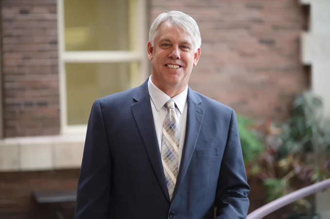 A professional portrait of an individual smiling in a business suit, standing in an indoor courtyard.