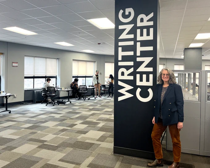 A woman leans against a sign that read "Writing Center". People sit at desks in the background.