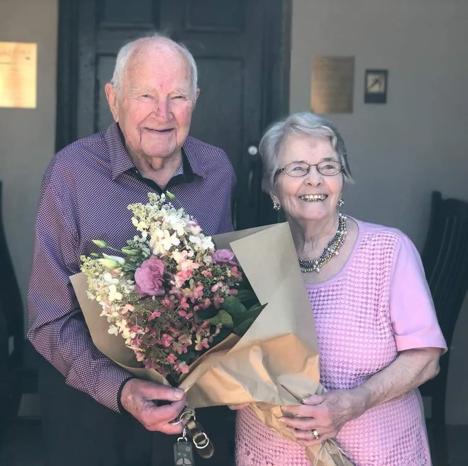 Harold and Margaret smiling together, with Harold holding a bouquet of flowers, standing in front of a doorway.