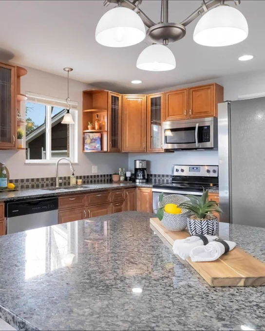Modern kitchen interior with stainless steel appliances, granite countertops, and wooden cabinets. Bright natural light streaming through a window by the sink area.