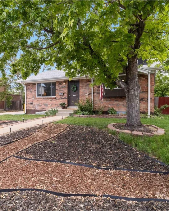 A charming brick suburban house adorned with an American flag, featuring a well-maintained lawn and landscaping under a large tree.