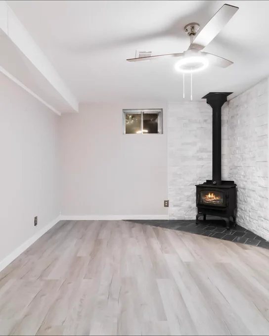 Interior view of a modern room featuring light hardwood floors, a white brick accent wall with a black wood-burning stove, and a ceiling fan. The room is well-lit and appears newly renovated.