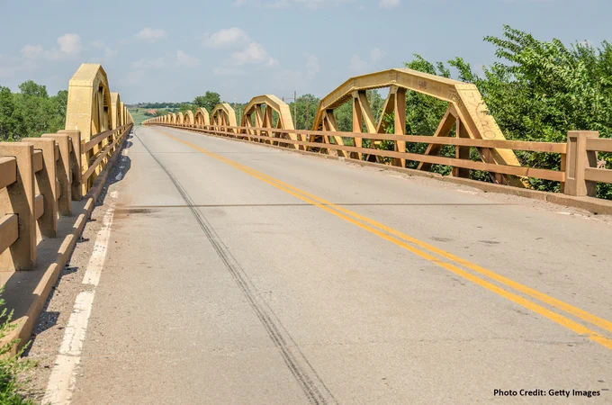 View of the historic Pony Truss bridge on Route 66 in Oklahoma, featuring a series of yellow arches and a clear sky. Farmland is seen in the background.
