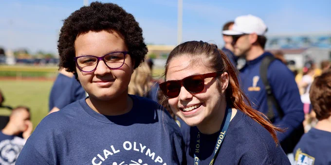 A smiling student and adult wearing blue Special Olympics shirts. It is a sunny day on an athletic field.