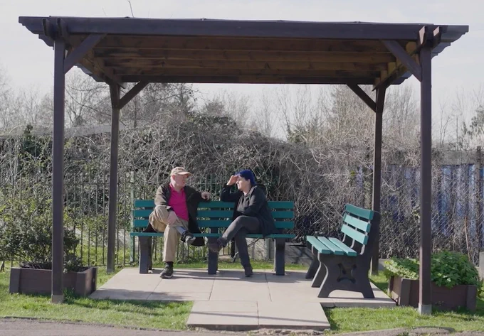 Two people sitting on a green bench underneath a pergola. 