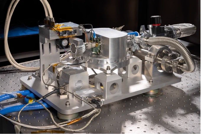 Close-up of a metallic scientific instrument with valves, pipes, and electronic components, set on a perforated lab table.